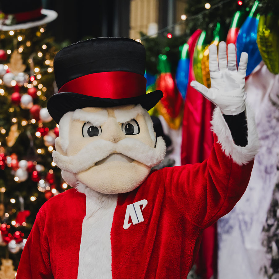 The Gov Mascot in a Santa suit waving at the camera while standing infront of a Christmas Tree.