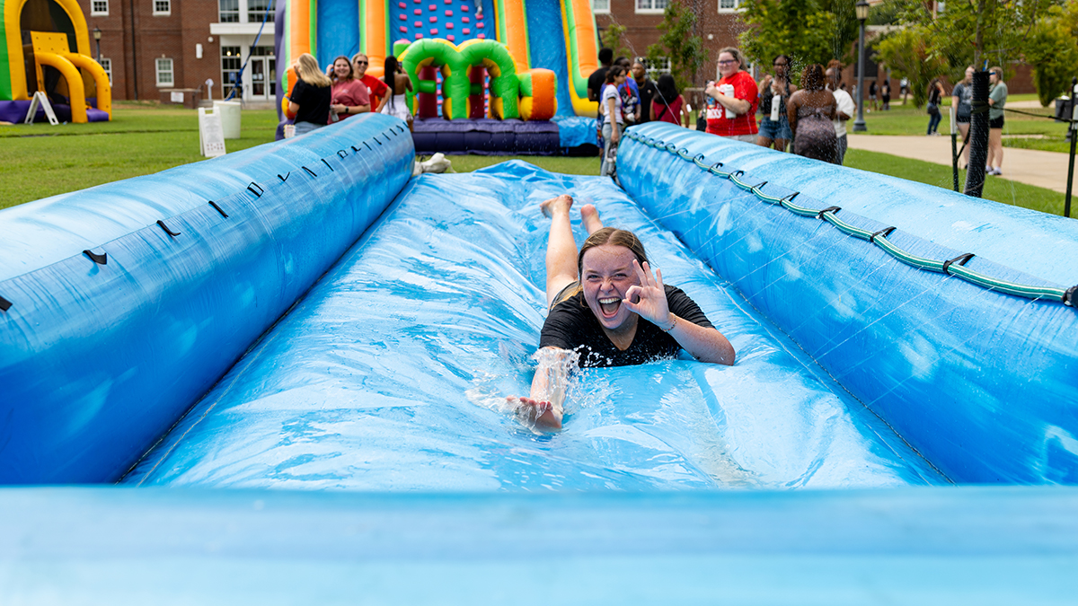 Female student sliding toward the camera on an inflateable slip-n-slide while holding up the Gov monocle hand signal.