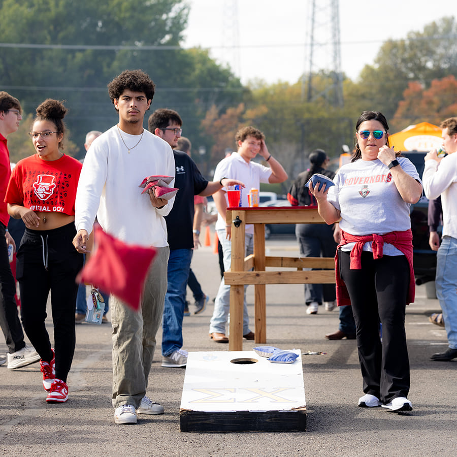 A mother and son throwing bags toward the camera during a game of cornhole in the parking lot while other students hang around in the background.