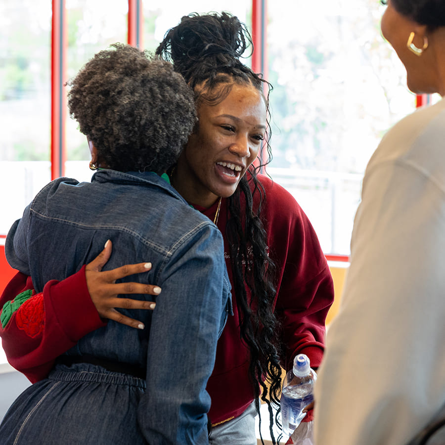 Woman in a maroon sweatshirt hugging a woman in a denim jacket indoors.