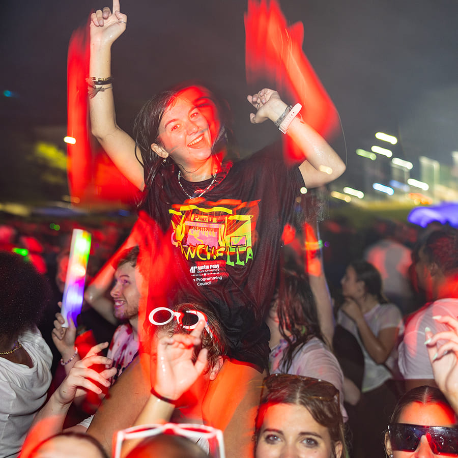Young woman on someone’s shoulders during Glowchella, crowded nighttime scene with red light trails