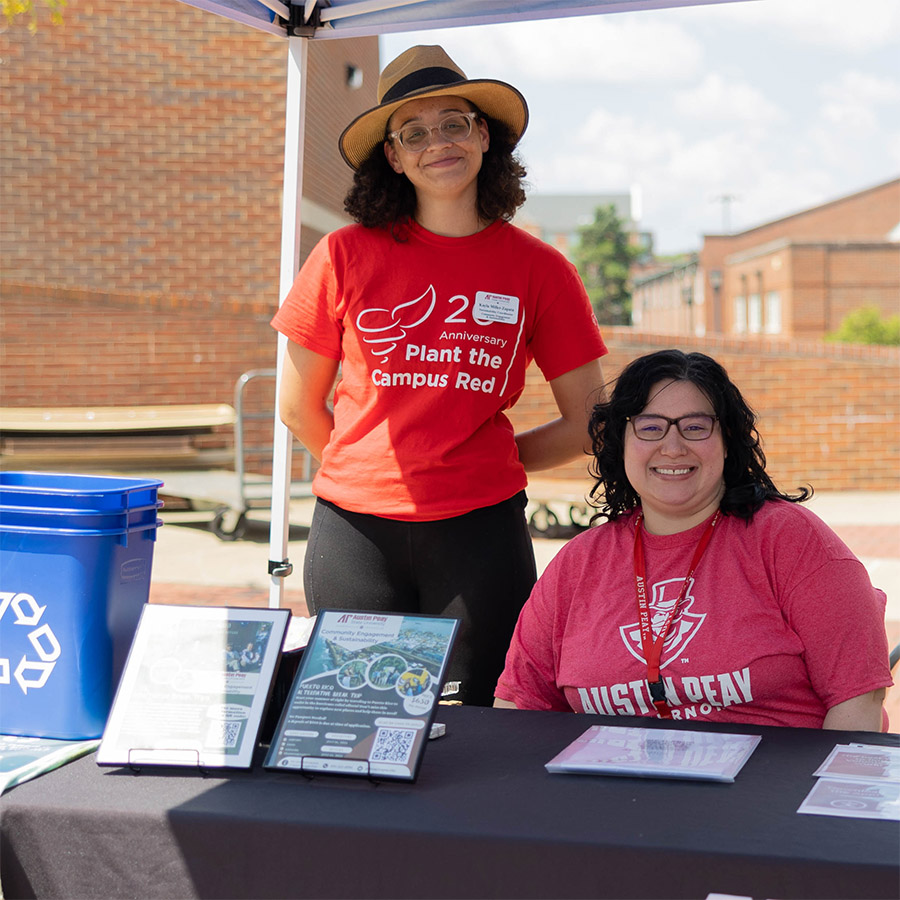 Two women at an Earth Day Fair table with informational materials and a recycling bin under a canopy.