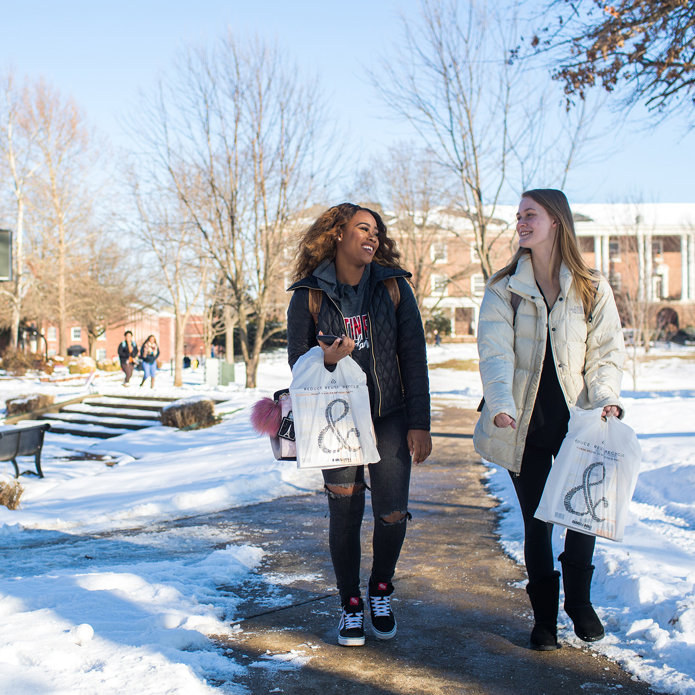Winter scene with snow on the ground showing two student walking with bags from the bookstore.