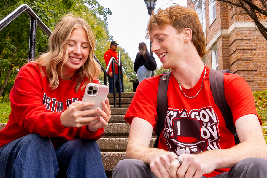 Two students sitting on stairs outside smiling and looking at a cellphone.