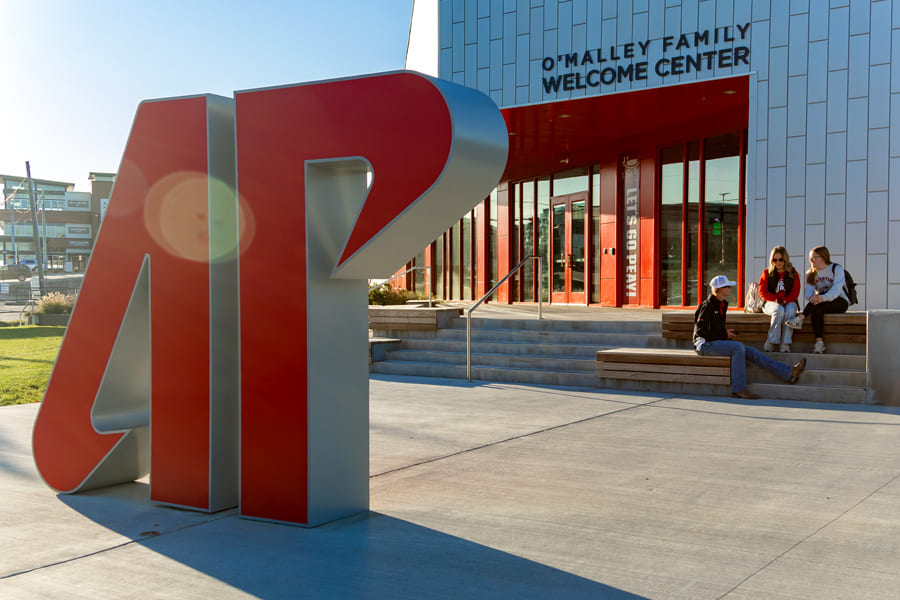 A small group of people sitting in front of the Welcome Center in the background and the giant 3d 'AP' sign in the foreground in front of the building with a lens flare and golden hour lighting.