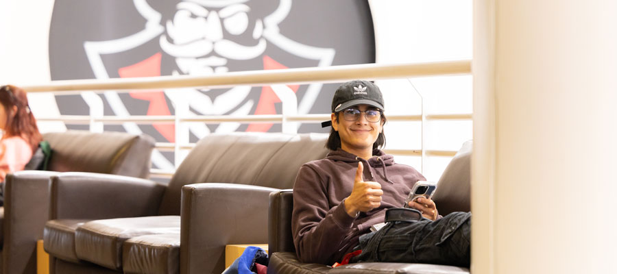 Student relaxing on a couch in the University Center smiling and giving the camera a thumbs up.