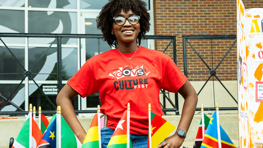 A student on the MUC plaza standing behind a table lined with small flags of several African countries— South Sudan, Ghana, Togo, Cameroon, DRC, Kenya—during the APSU Culture Fest event.