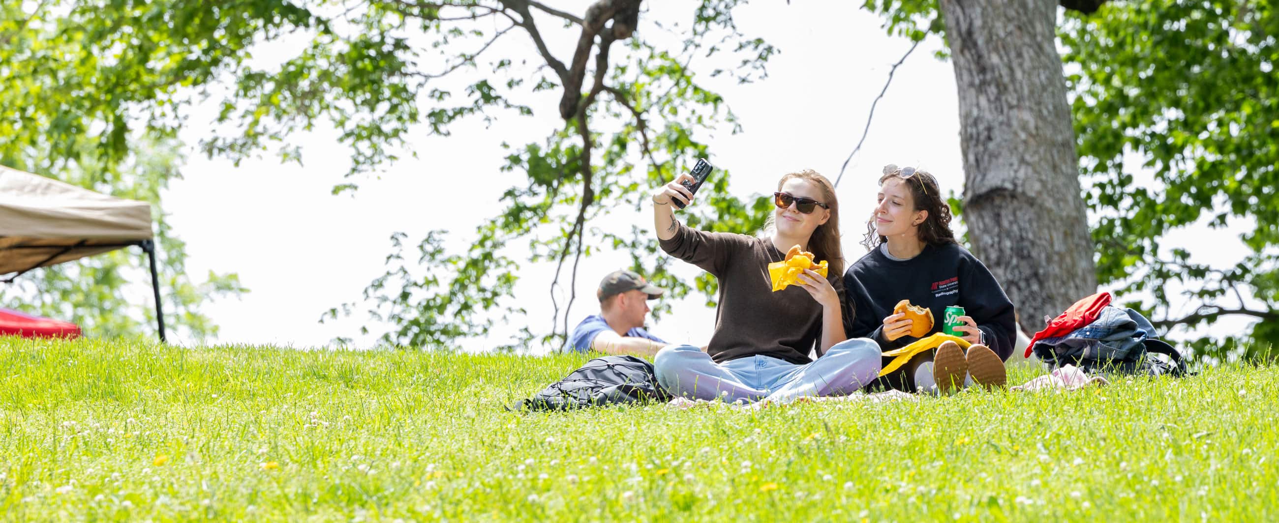 Student taking a selfie while eating cheeseburgers while sitting on the grass during Study on the Hill. 