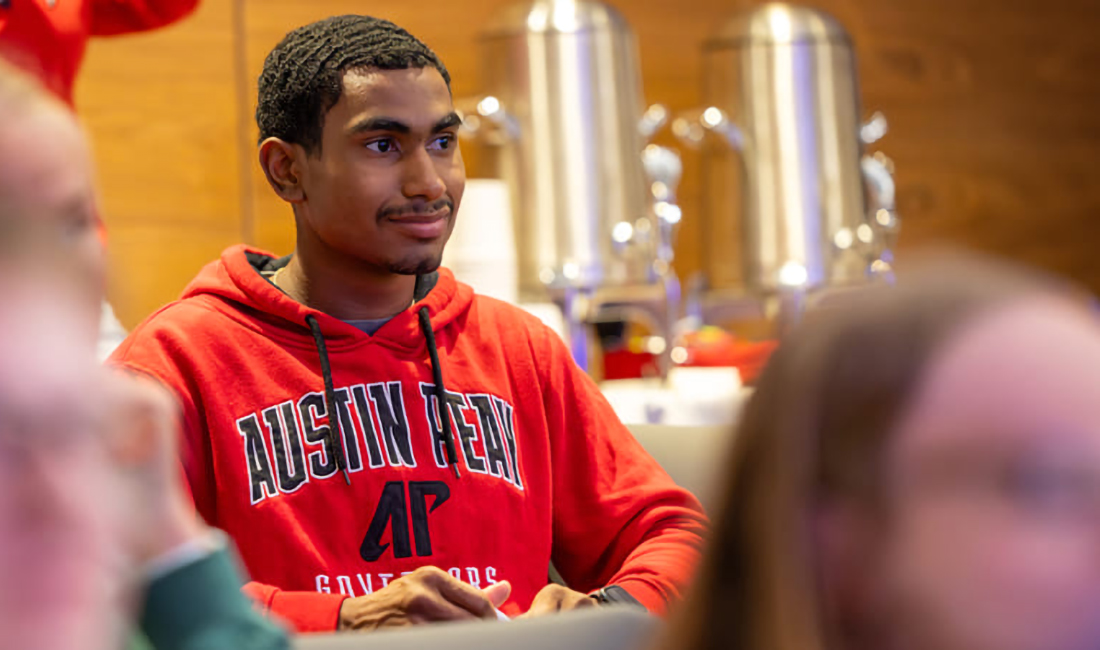 A student sitting between two other out-of-focus people in the foreground, listening intently during an event and smiling.