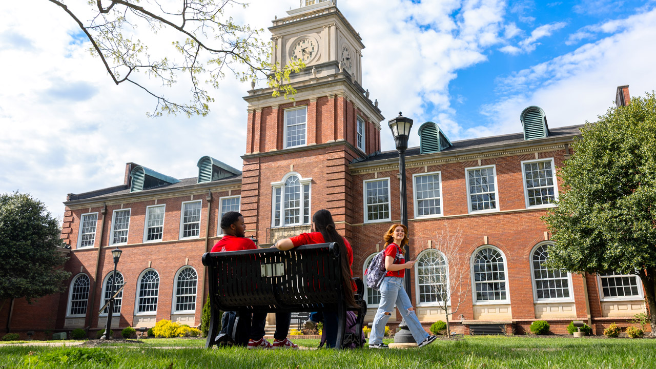 Students sitting, talking on a bench in front of the Browning building while another student walks by.