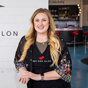 Diane standing in her salon wearing a black apron with the 'Red Bird Salon' logo on it.