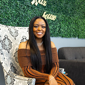 Jasmine sitting in an armchair in front of a faux foliage wall with the 'Wax House' logo.