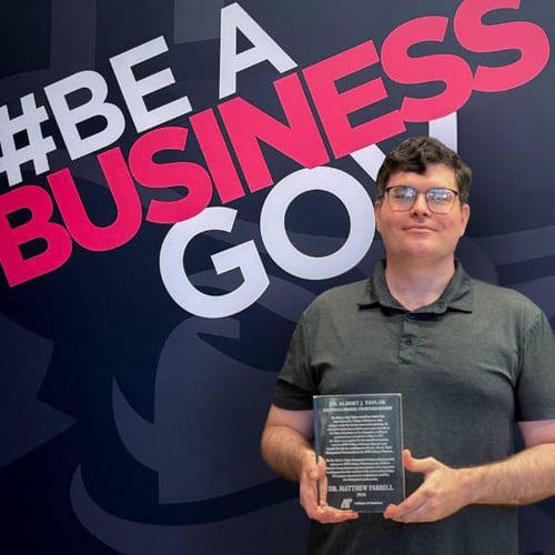 Dr. Matthew Farrell holding his award in front of a backdrop that reads "#Be A Business Gov."