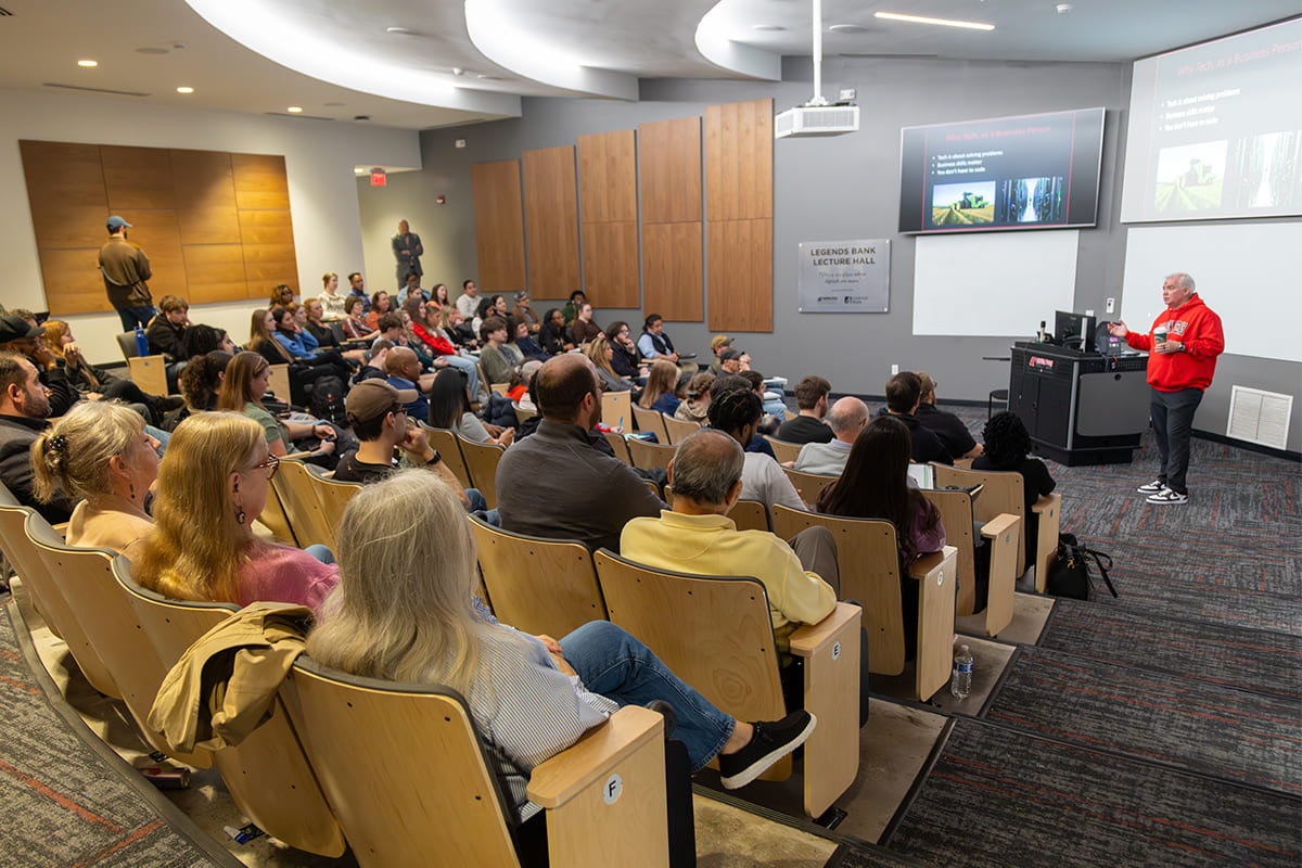 Presenter in a red hoodie speaking to a seated audience in Legends Bank Lecture Hall