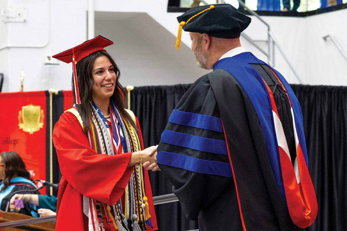 Daniela in her graduation regalia with numerous cords draped around her neck, shaking Dr. Hepner's hand as she walks across the ceremony stage.