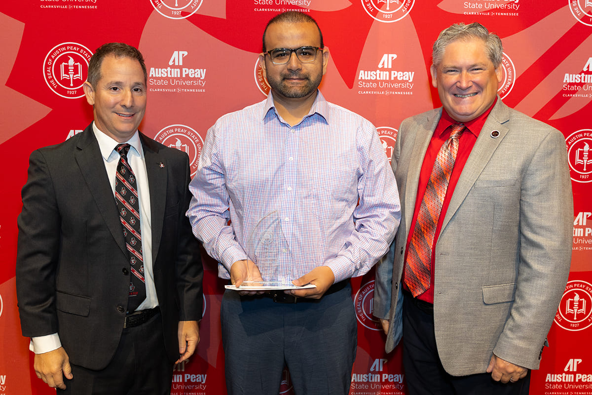 Dr. Cifci holding his award while posing with the Provost and President in front of a red step-and-repeat.