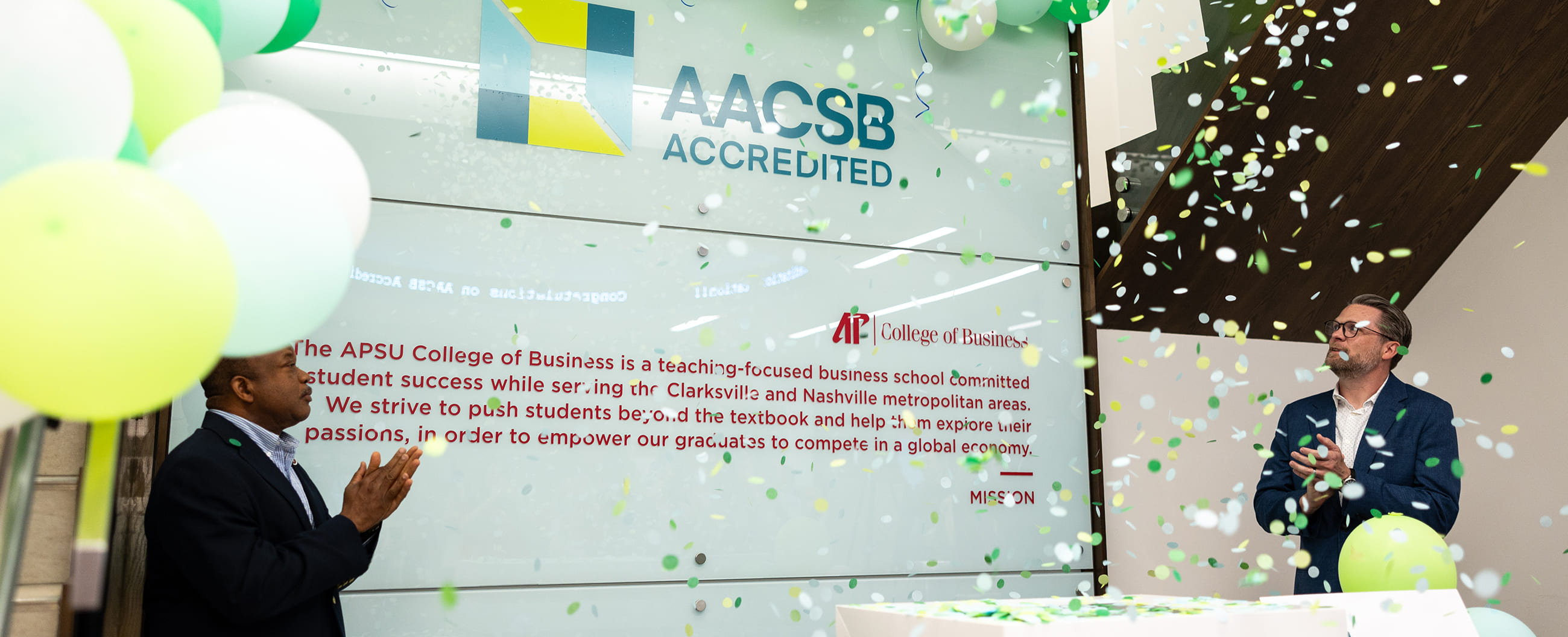 AACSB Accredited wall display at APSU College of Business with two men clapping amid confetti and balloons.