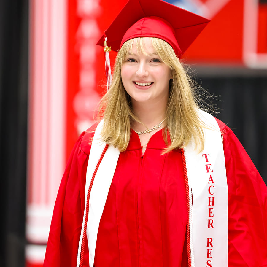 Smiling graduate in a red regalia wearing a white stole labeled “Teacher Res-" (resident)