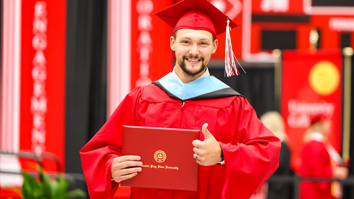 Image of a male graduate in red regalia smiling at the camera while holding up his diploma and a thumbs up.