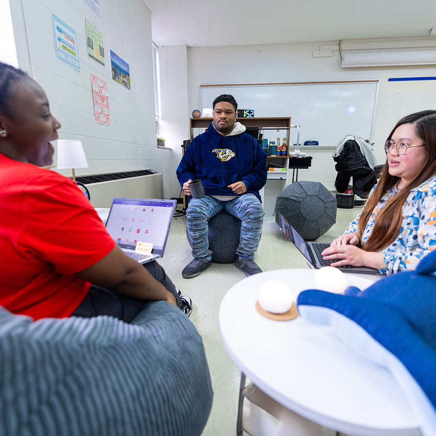 Three students sitting in a circle with laptops having an enjoyable discussion in a classroom setting..