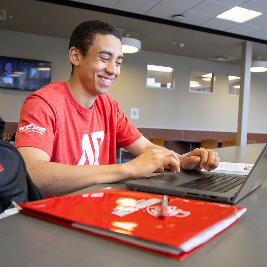 Student sitting in front of his laptop smiling with a bright red folder sitting on the table beside him in the foreground.