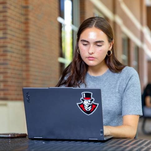 Young woman using a laptop at an outdoor table beside a brick building.