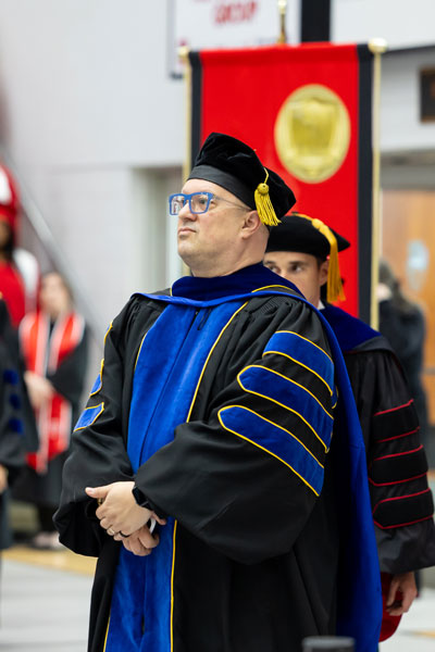 Faculty member in black regalia with blue accents walking into commencement.