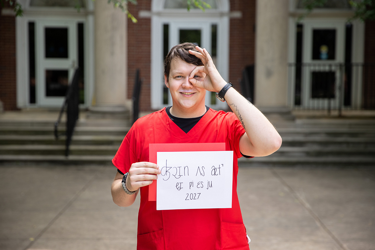Student in red scrubs holding the monocle sign (ok symbol) over his left eye and a handwritten sign in the other hand that reads "Join Us At APSU 2027" in the International Phonetic Alphabet.