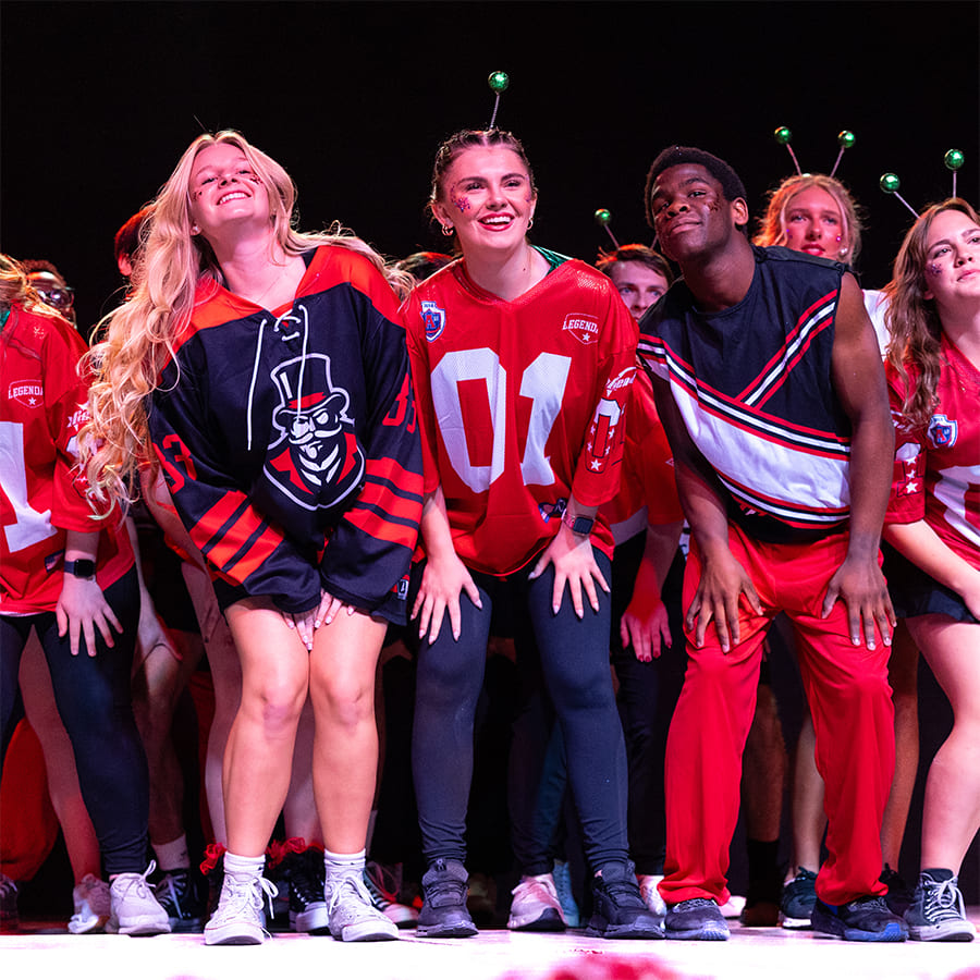 Students perform in a lip sync battle on stage, wearing red, black, and white APSU jerseys/outfits and showcasing enthusiastic expressions.