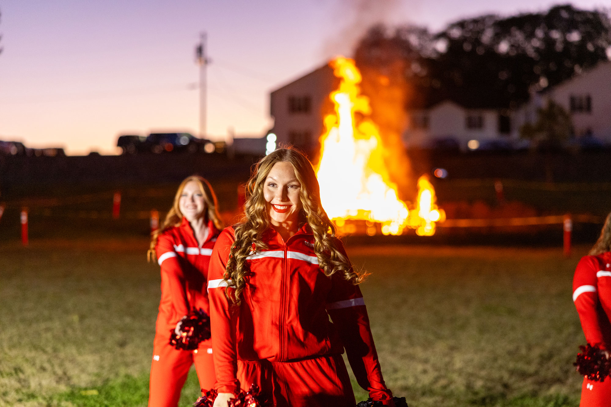 Cheerleaders in red jumpsuits performing a routine in front of a giant bonfire. 