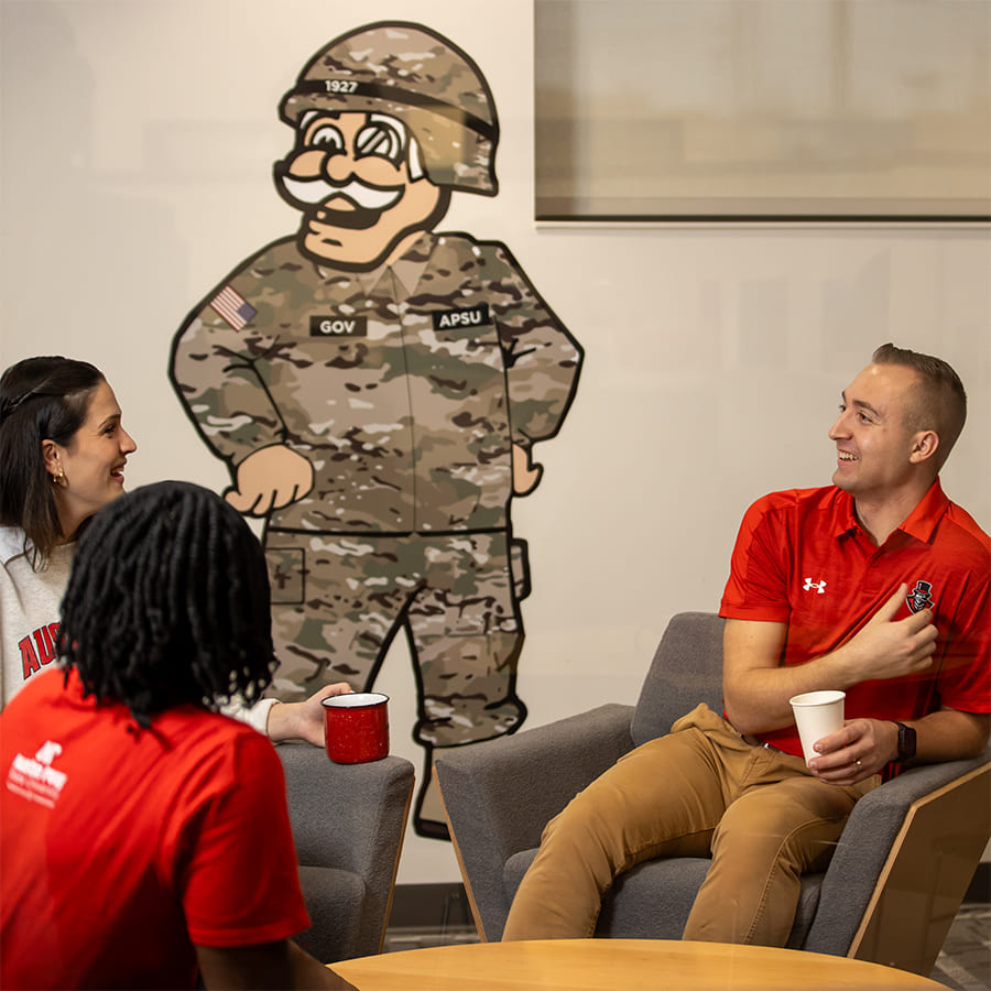 Military students in civilian clothing sitting in a circle of arm chairs chatting and hanging out with the illustration of the Governor mascot in an army uniform on the wall behind them.