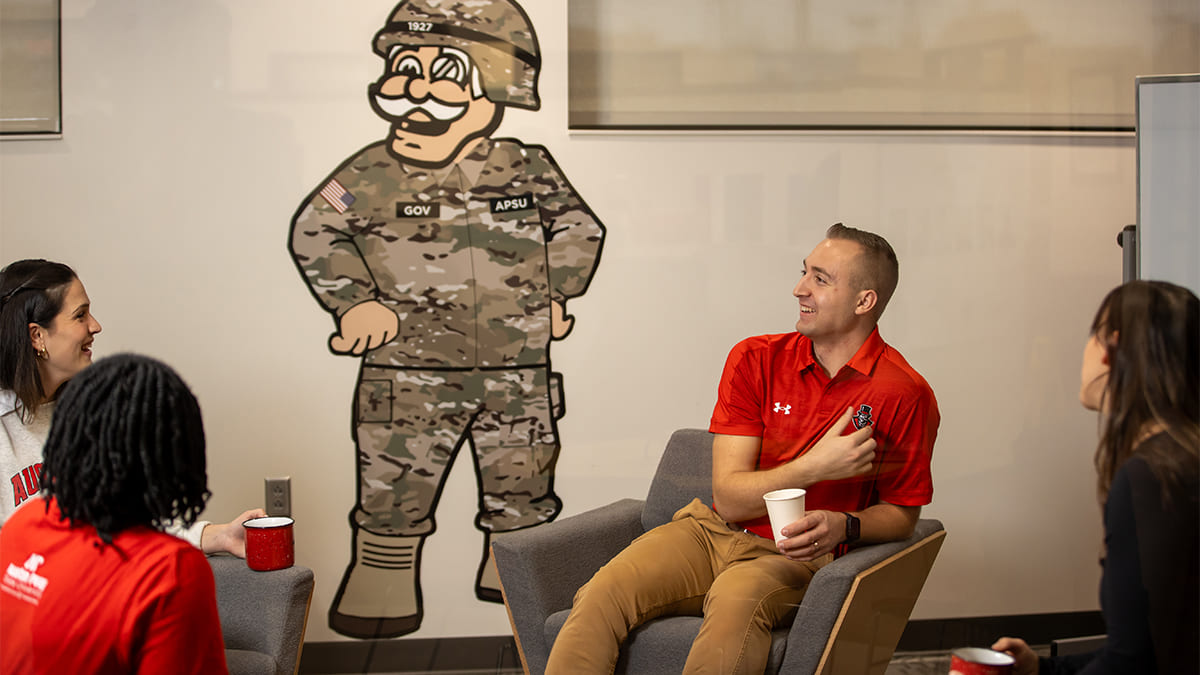 Military students in civilian clothing sitting in a circle of arm chairs chatting and hanging out with the illustration of the Governor mascot in an army uniform on the wall behind them.