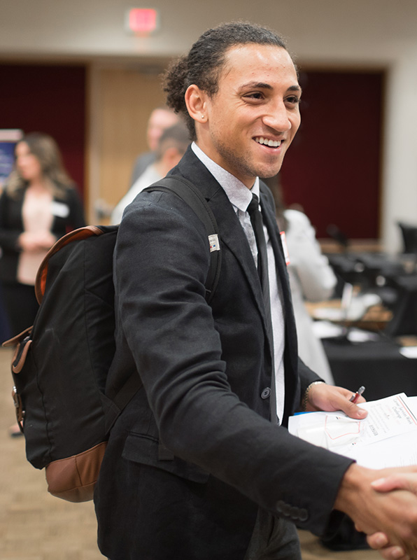 Male student wearing a black suit and backpack shaking hands at a career fair.