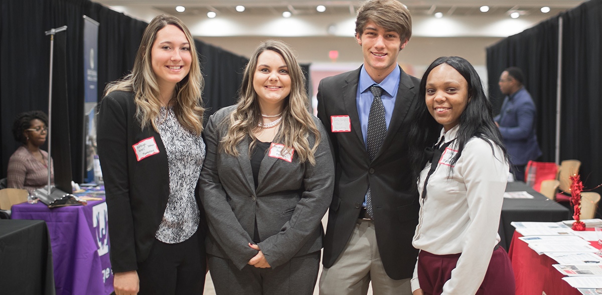 A group of four students in professional clothing posing for the camera at a career fair.