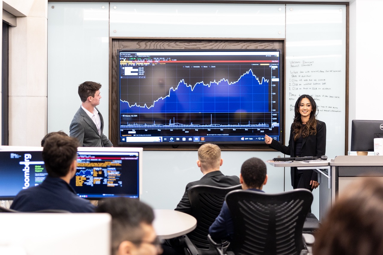 In a modern classroom or business setting, a woman in a dark blazer stands at the front, smiling and gesturing toward a large digital screen. The screen displays a financial line graph, similar to a stock chart. To her left, a man in a gray suit jacket also looks at the screen. In the foreground, the backs of several students or attendees are visible as they sit at tables, focusing on the presentation. A computer monitor in the lower-left corner shows a Bloomberg terminal interface.