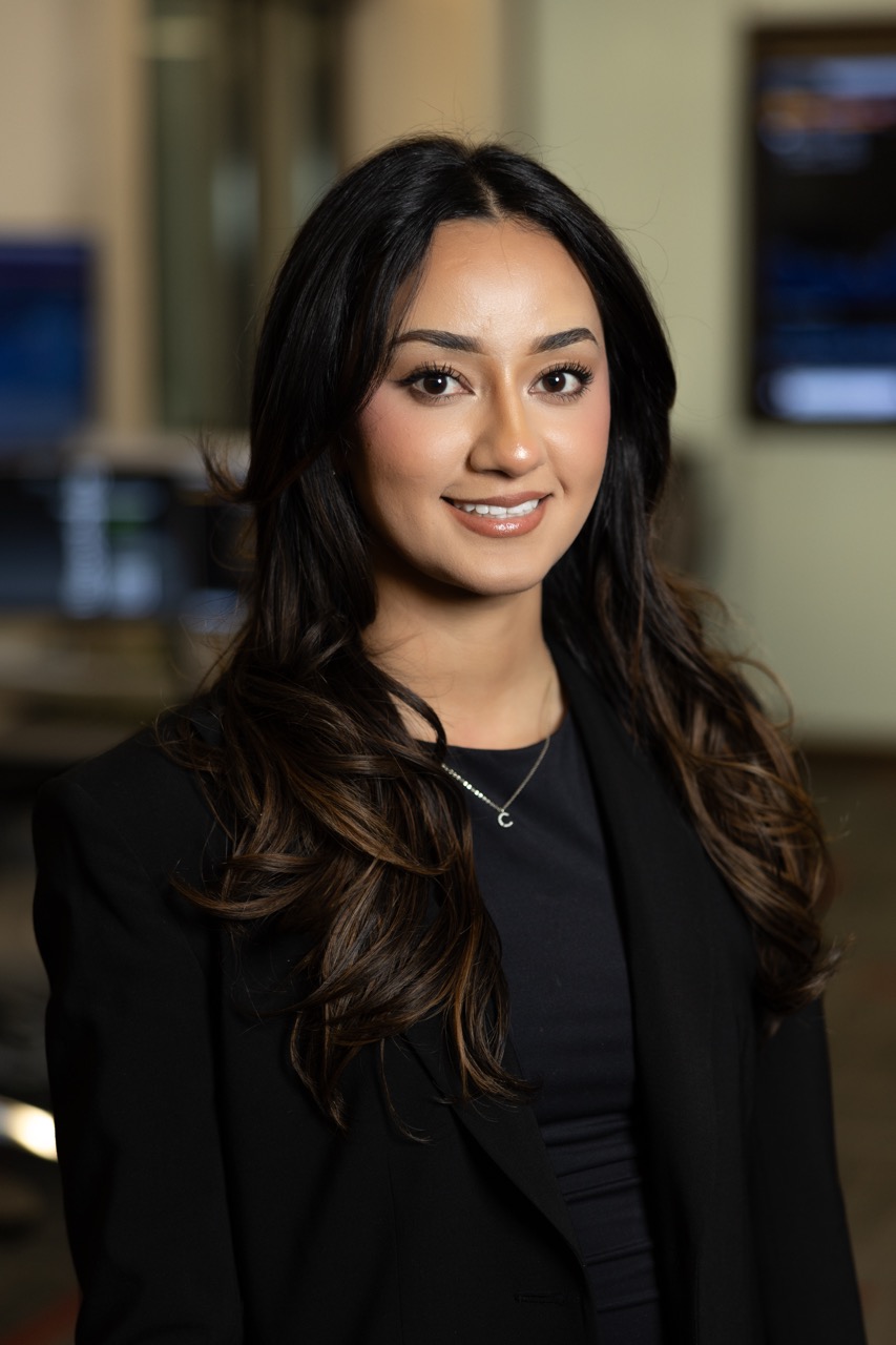 This is a medium, chest-up portrait of a young woman smiling warmly at the camera. She has long, dark brown hair with lighter highlights, styled in waves. She is wearing a black blazer over a black top and a simple silver necklace. The background is blurred but appears to be an indoor office or lab setting.