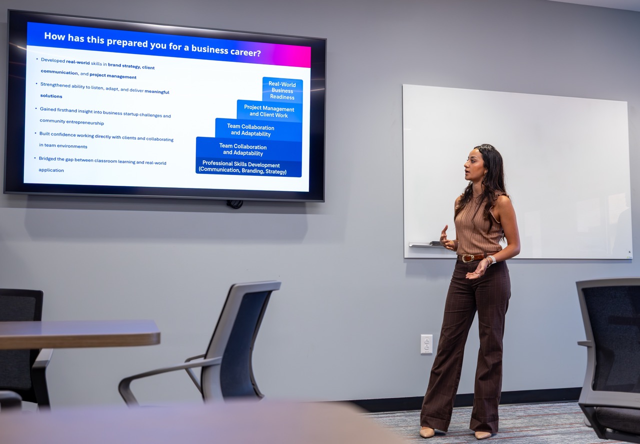 In a modern conference room, a woman stands giving a presentation. She is wearing a brown sleeveless top and dark trousers, gesturing with her hands as she speaks. Beside her, a large digital screen displays a slide titled, "How has this prepared you for a business career?" The slide features a bulleted list on the left and a blue stacked diagram on the right, detailing skills like "Project Management" and "Team Collaboration." A white-erase board is visible on the wall behind her. The foreground shows the blurred tops of conference tables and chairs.