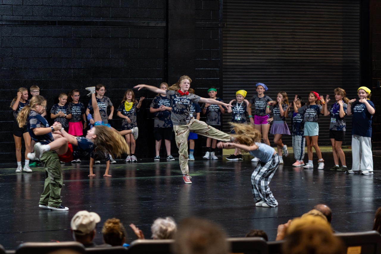 A group of children performing a dynamic dance routine on a black stage, featuring a girl in a red bandana doing a high kick.