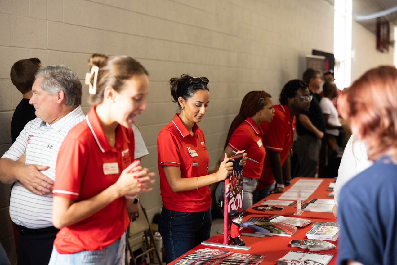 This photo captures a busy scene at what appears to be a college fair or event. In the foreground, several people, including a young woman smiling brightly on the left and another woman beside her, stand behind a long table covered with a red cloth. They are all wearing matching red polo shirts with a logo. The woman in the center is focused on adjusting a small, vertical banner on the table that says "GOVS." The table is neatly arranged with brochures, pamphlets, and other promotional materials. In the background, more people, both attendees and staff, are lined up along the table and milling about, indicating an active event.