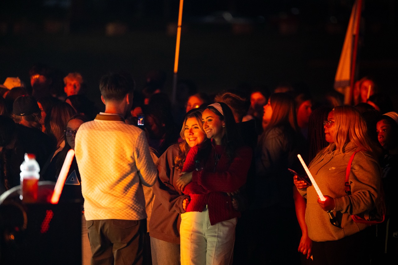 This photo captures a nighttime outdoor gathering, likely a bonfire or pep rally, illuminated by warm light. In the center, two young women are posing closely together for a photo, smiling brightly. The woman on the right, wearing a white headband and red cardigan, has her arm around the woman in the brown sweatshirt next to her. A young man, seen from behind, stands just in front of them, holding up a smartphone to take their picture. The surrounding crowd is bathed in the glow of the fire, which is off-camera. Several people, including a woman on the far right, are holding white or orange glow sticks, adding to the festive, dark ambiance.
