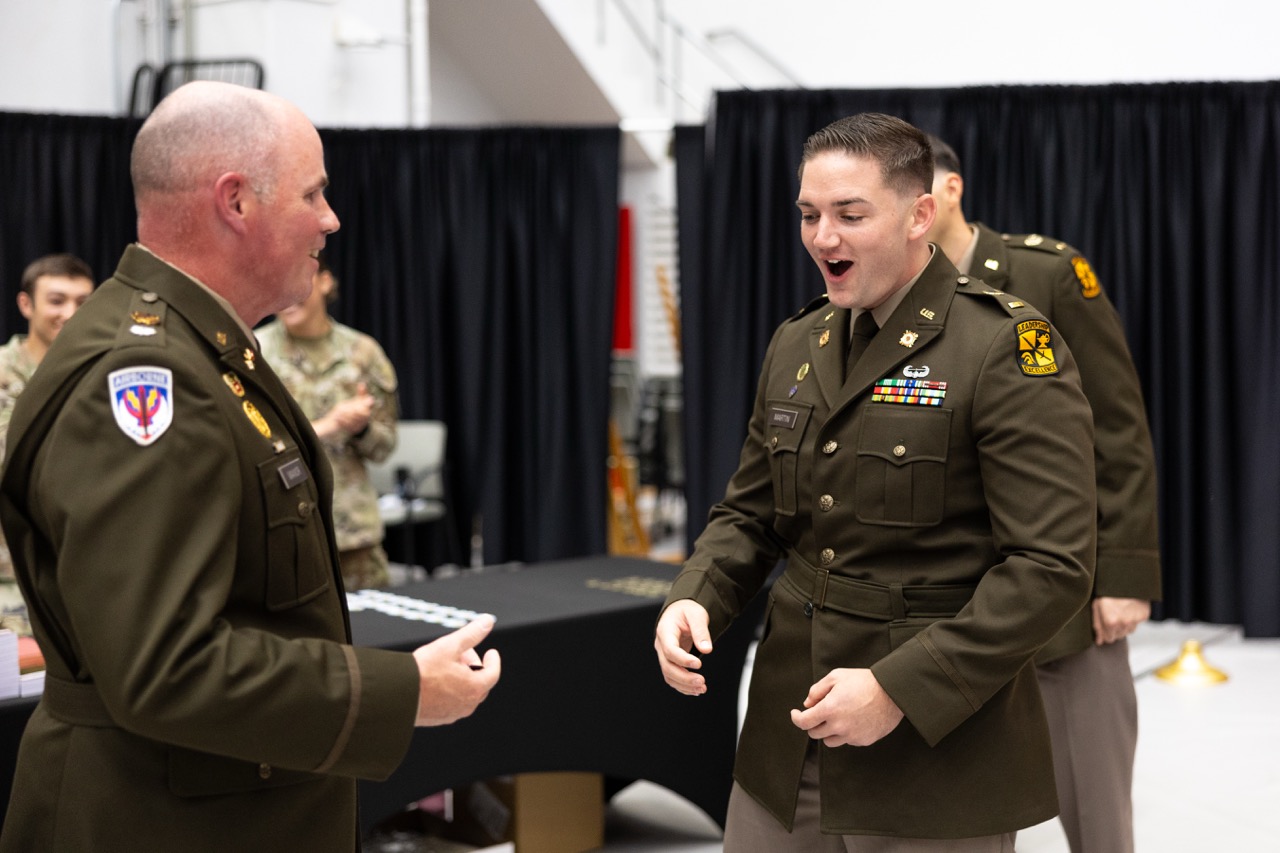 A candid shot of the newly commissioned second lieutenant in his green Army dress uniform standing across from a higher-ranking officer. The young lieutenant is looking down with his mouth open in an expression of happy surprise and excitement, while the older officer looks on with a smile. Black curtains are visible in the background.
