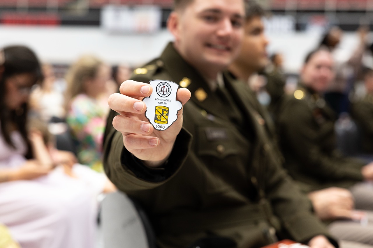 A close-up of a hand holding up a custom, shield-shaped challenge coin featuring a top hat graphic. The coin reads "Governor's Guard," "Leadership Excellence," and "1000." In the softly blurred background, the same young soldier in his green dress uniform sits in the audience and smiles proudly.
