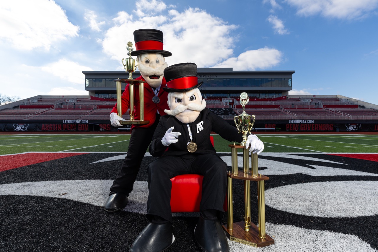 Two Austin Peay Governor mascots posing on a football field with large gold championship trophies. One mascot sits in a red chair while the other stands behind; both are dressed in their signature red and black formal attire with top hats.