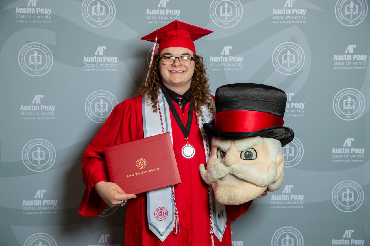 A smiling graduate with long curly hair and glasses wears a red cap and gown. They are posing in front of a grey Austin Peay State University backdrop while holding a red diploma cover and the Governor mascot head.