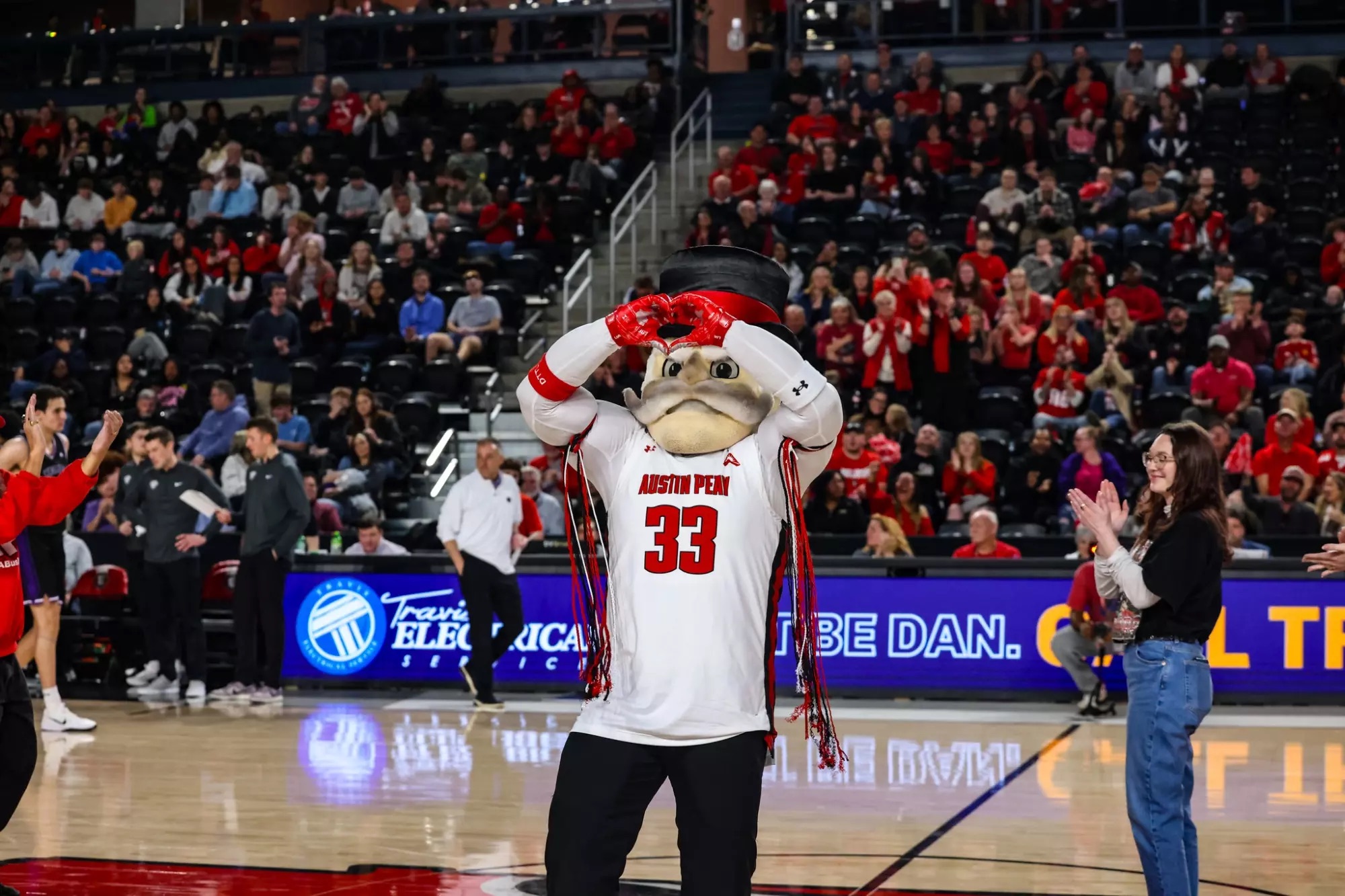 A graduate in red commencement attire sits on a black metal park bench in an outdoor setting. He rests his hand on the top hat of the Governor mascot head, which is sitting on the bench beside him.