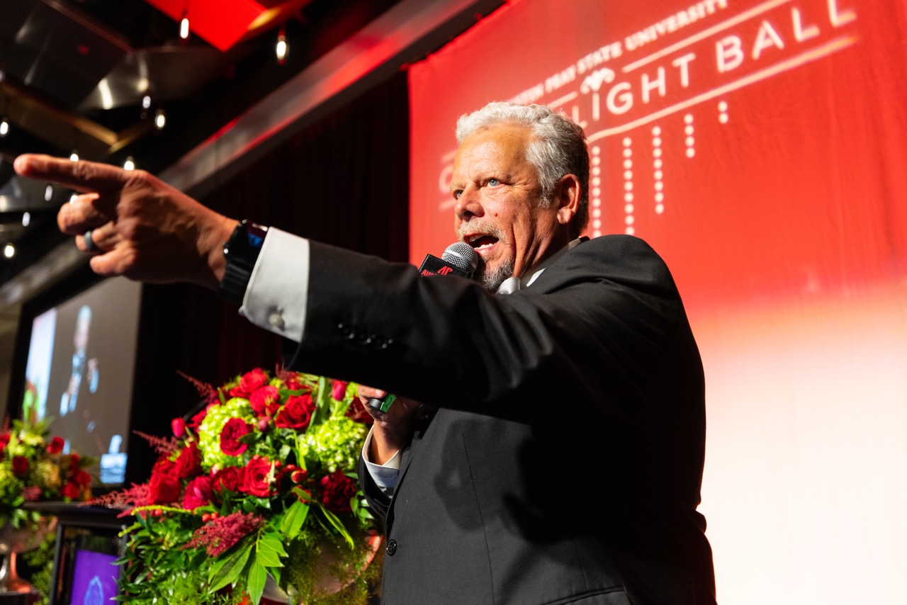 A man in a formal black tuxedo and white bow tie speaks into a microphone while pointing emphatically off-camera. He stands on a stage decorated with vibrant red roses and green hydrangeas, with a large red banner in the background reading "Austin Peay State University Candlelight Ball."