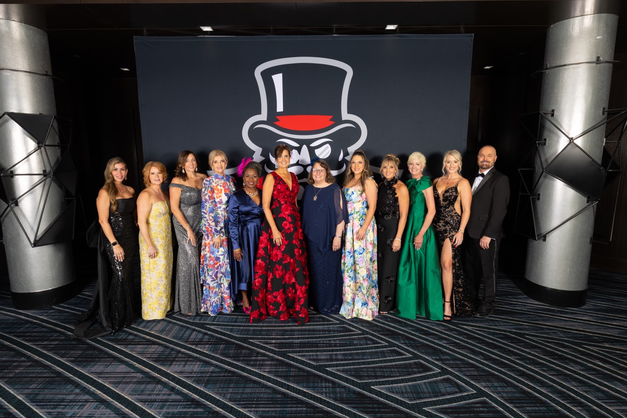 A large group of guests in elegant formal attire, including sequined gowns and tuxedos, poses for a group photo in a modern lobby. They are positioned in front of a dark backdrop featuring a large white and red mascot logo of a man in a top hat.