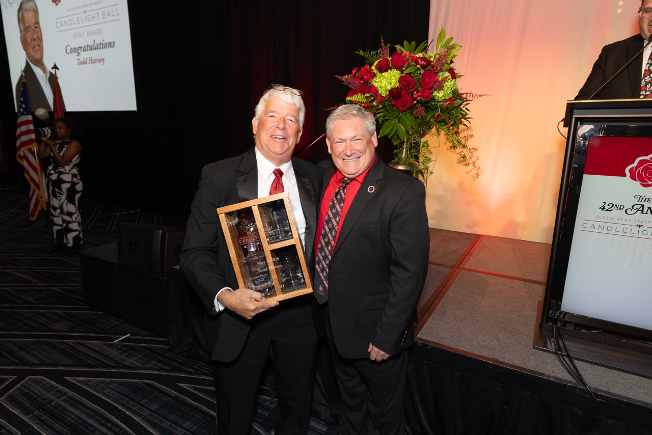 Two men in suits stand together on a stage smiling for a photo. The man on the left holds a wooden display case containing etched glass decanters and glasses. A screen in the background displays a "Spirit Award" congratulating Todd Harvey.