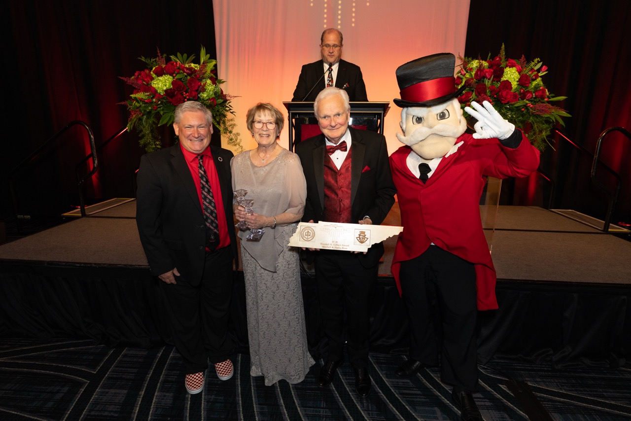 A group of four stands on a stage during an awards ceremony. A woman in a silver lace gown holds a glass trophy, while a man in a tuxedo holds a large award shaped like the state of Tennessee. They are joined by a man in a suit and the university mascot, Governor Peay, in his signature red coat and top hat.