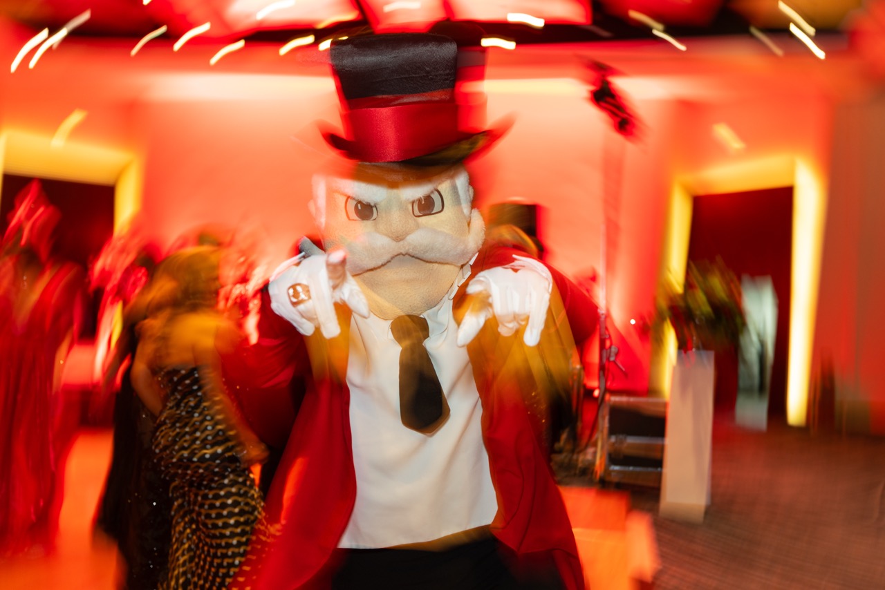 A dynamic, slightly blurred action shot of the Governor Peay mascot on a dance floor. The mascot, wearing a red coat and black top hat, is pointing both gloved hands directly at the camera while guests dance in the background under warm red lighting.